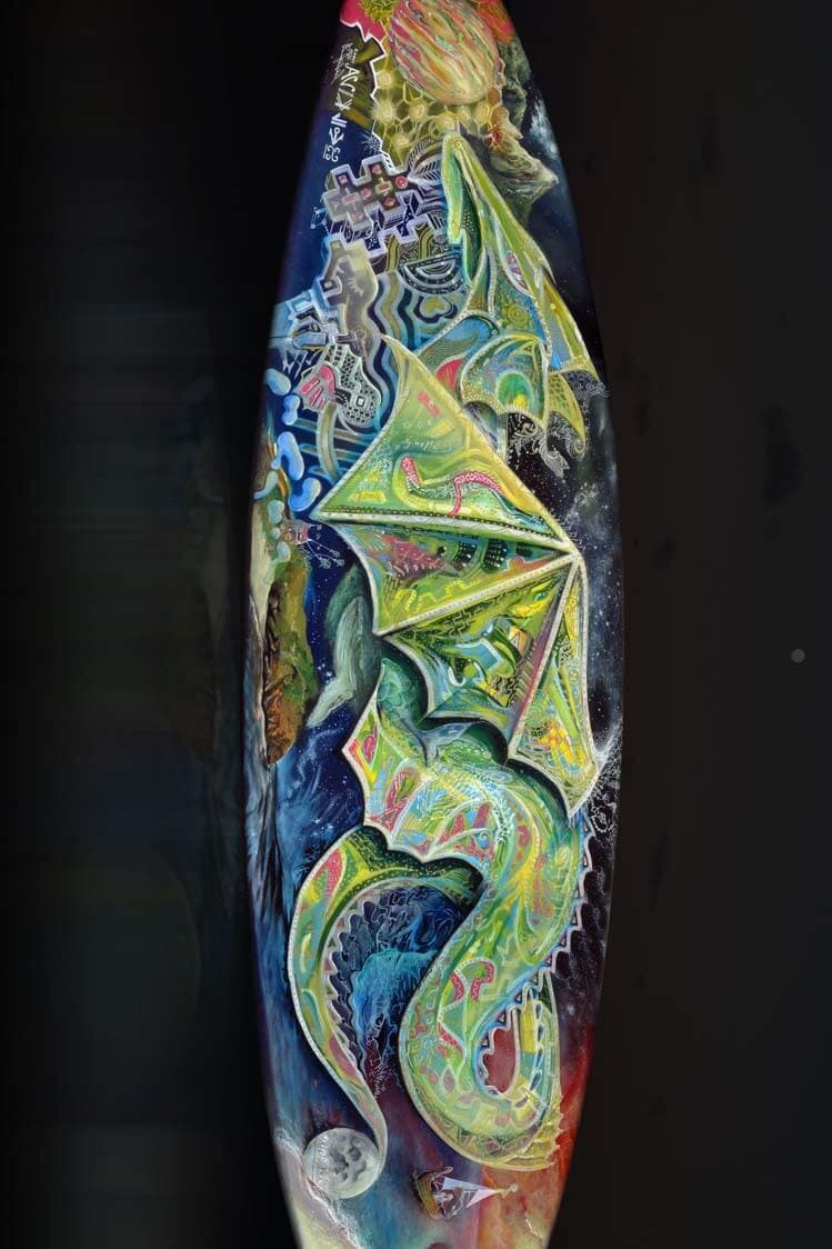 A surfboard painted end to end with a winged green dragon in Posca and paint, set among cosmic patterns, a moon and dense graphic detail.
