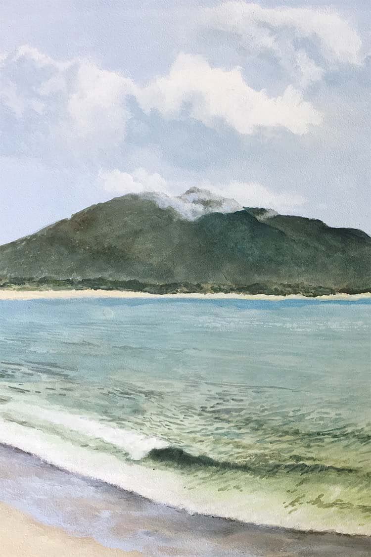 A green mountain rising across a calm bay, cloud resting on its summit, with a soft wave breaking on pale sand in the foreground.