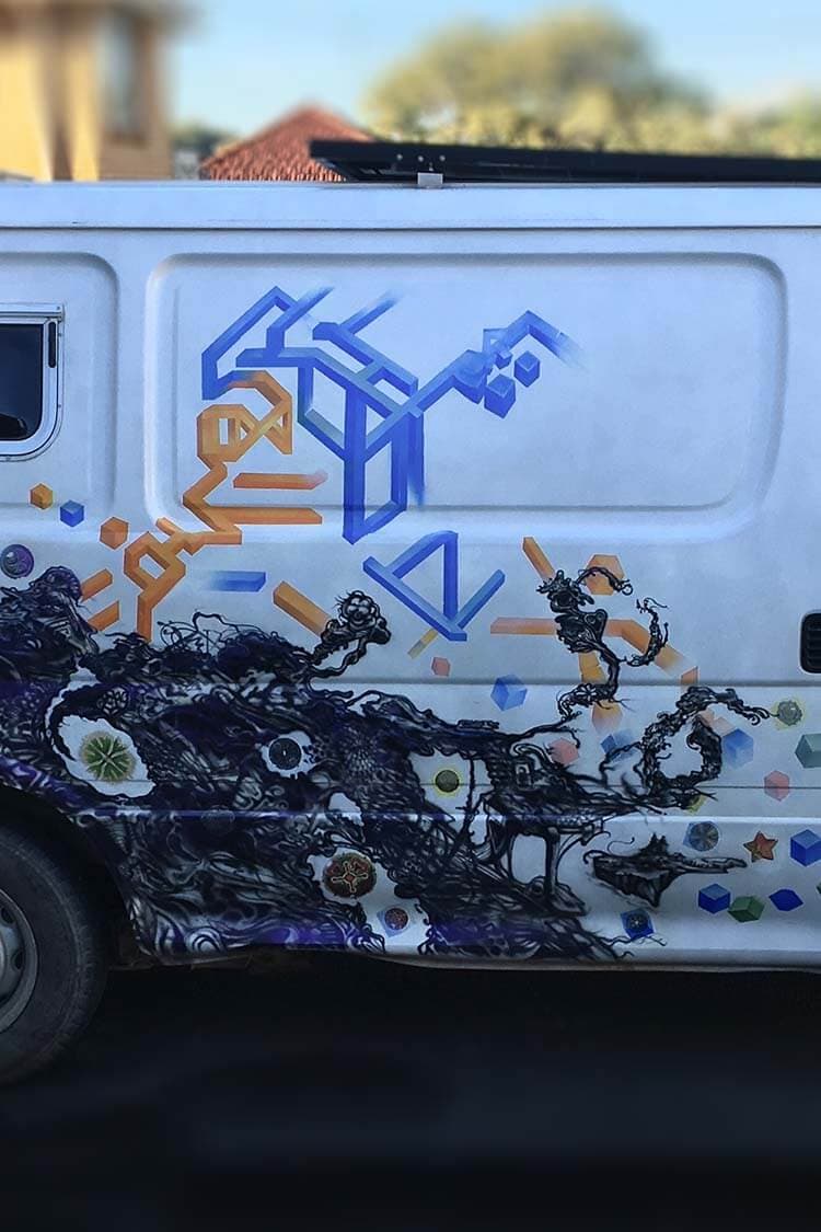 A white van painted along its side with blue isometric line forms, orange geometric shapes and dark, tangled botanical ink work studded with flowers and coloured cubes.